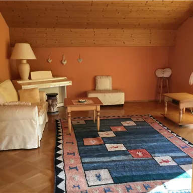 A cozy living room with orange walls and a wooden beam ceiling. There is a comfortable sofa, a coffee table, and a colorful rug.