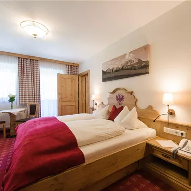 A cozy bedroom with a double bed and red accents. Windows with plenty of natural light and a small table with chairs in the background.