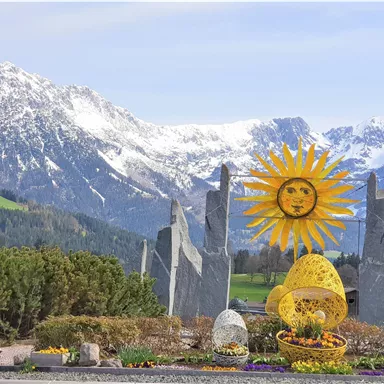 Eine bunte Sonnenskulptur steht vor schneebedeckten Bergen. Umgeben von Blumen und grünen Büschen vermittelt die Szene ein fröhliches Ambiente.
