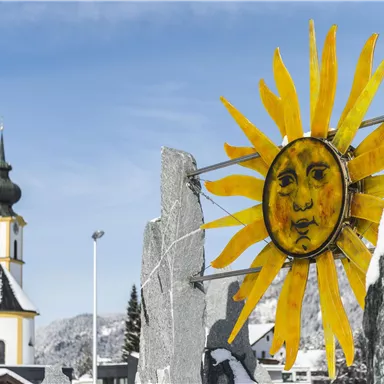 A sunny artwork with a smiling face stands in front of a snowy landscape. In the background, a church with a green tower can be seen.