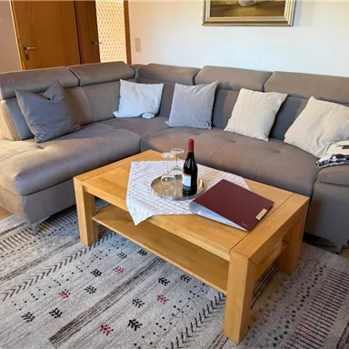 A cozy living room with a U-shaped sofa and many cushions. On the table, there is a bottle of wine, a glass, and a tablecloth.