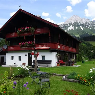 A traditional Alpine house with a red facade and flower boxes. In the background, majestic mountains and a green meadow can be seen.