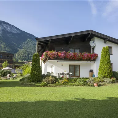 Ein charmantes Haus mit blühenden Fensterkästen und einem gepflegten Garten. Im Hintergrund sind grüne Berge zu sehen.