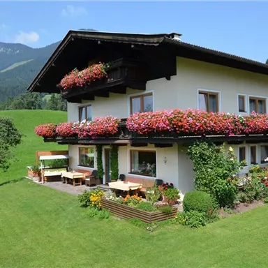 Ein charmantes Haus mit blühenden Balkonblumen in einer grünen Umgebung. Im Hintergrund sind sanfte Hügel und Berge sichtbar.