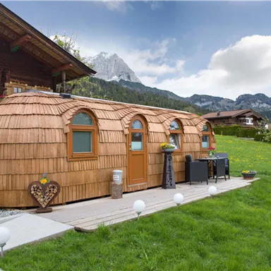 Ein modernes Holzhaus mit einem einzigartigen, runden Design steht in einer grünen Umgebung. Im Hintergrund sind malerische Berge und ein klarer Himmel zu sehen.
