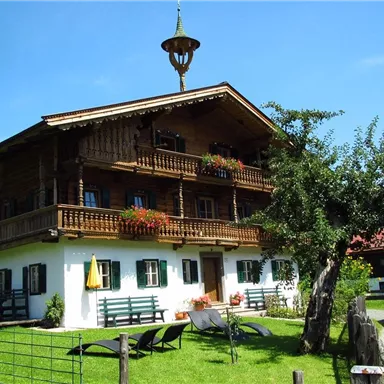 Ein traditionelles alpenländisches Holzhaus mit einem Balkon und bunten Blumen. Im Vordergrund gibt es eine grüne Wiese und einige Möbel zum Entspannen.