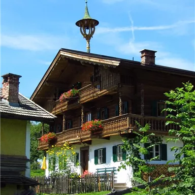 Ein traditionelles alpenländisches Haus mit einem markanten Wetterhahn. Es ist von grünen Wiesen und bunten Blumen umgeben.