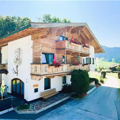 A charming wooden house with several balconies and colorful flower plantings.
Surrounded by green countryside and clear sky.