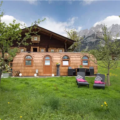 Eine gemütliche Holzhütte im Alpenstil umgeben von grünen Wiesen und Bergen. Vor der Hütte befinden sich Liegestühle und ein schöner Garten.