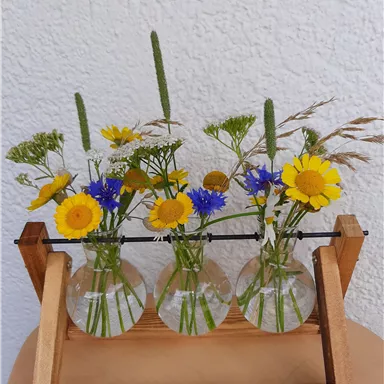 A wooden vase with three glass tubes, filled with colorful flowers. The flowers include yellow, blue, and green blooms, giving the arrangement a fresh touch.