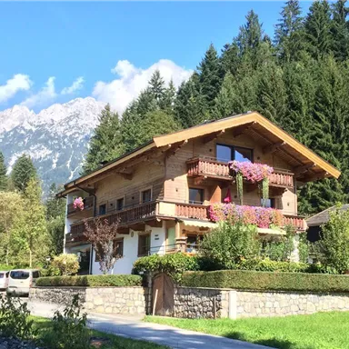 A beautiful wooden house with balconies and colorful flowers. In the background, high mountains and a clear blue sky are visible.