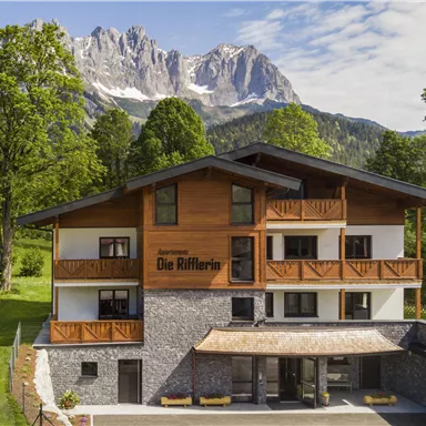 Ein modernes Gebäude mit einem Holzfassade, umgeben von grüner Wiese und Bäumen. Im Hintergrund sind majestätische Berge zu sehen.