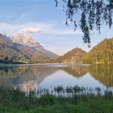 Ein ruhiger See umgeben von hohen Bergen und dichten Wäldern. Der Himmel ist blau mit wenigen Wolken.