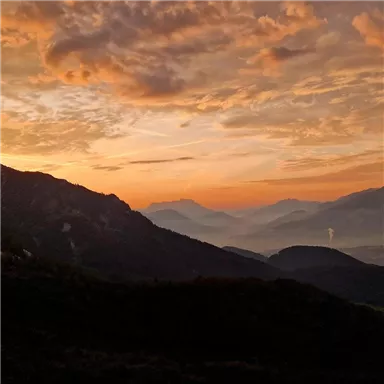 Ein atemberaubender Sonnenuntergang über den Bergen, mit orangefarbenen und violetten Wolken. Die Landschaft zeigt sanfte Hügel und eine mystische Atmosphäre.