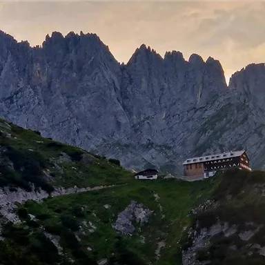 Eine beeindruckende Berglandschaft mit steilen Felsen und dramatischem Himmel. Am Hang steht ein traditionelles Almhüttengebäude.