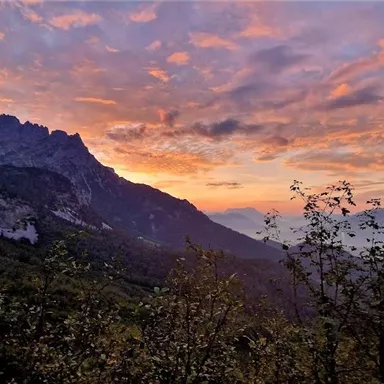 Eine beeindruckende Berglandschaft bei Sonnenuntergang mit bunten Wolken am Himmel. Bäume und Hügel im Vordergrund runden das friedliche Naturbild ab.