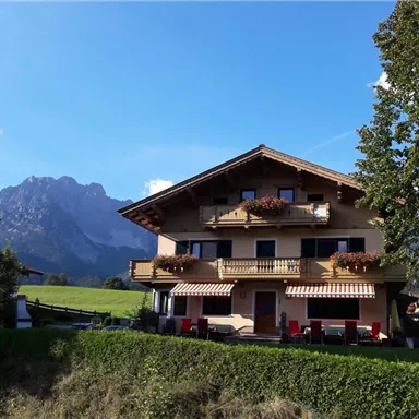 A charming mountain house with a balcony and flowers. Majestic mountains rise in the background under a clear blue sky.