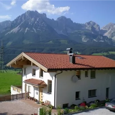 Ein schönes Haus mit rotem Dach in einer grünen Landschaft. Im Hintergrund erheben sich majestätische Berge unter einem blauen Himmel.