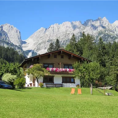 Ein hübsches Haus in den Bergen, umgeben von Bäumen und Wiesen. Im Hintergrund erheben sich majestätische Berggipfel unter einem klaren blauen Himmel.