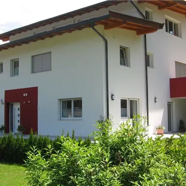 A modern, two-story house with a white facade and a red accent. Surrounded by green bushes and trees.