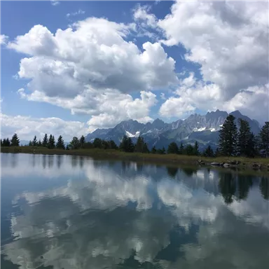 A peaceful landscape with a clear lake and majestic mountains in the background. The clouds gently reflect in the water.