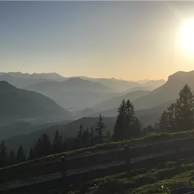 A picturesque mountain landscape with gentle hills and a radiant sunset. In the foreground, trees and a wooden railing are visible.