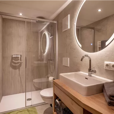 Ein modernes Badezimmer mit einer Glasdusche und einem eleganten Waschbecken. Die Wände sind in neutralen Farben gehalten, begleitet von einer runden, beleuchteten Spiegelfläche.