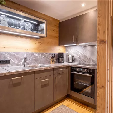 Eine moderne Küche mit grauen Schränken und einer Marmoroberfläche. Im Hintergrund sind ein Fernseher und eine Holzdecke sichtbar.