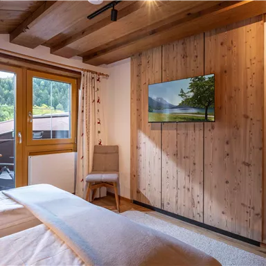 Ein helles Schlafzimmer mit Holzdecke und einem großen Fenster. An der Wand hängt ein Fernseher und ein Stuhl steht neben dem Bett.