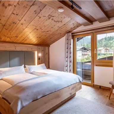 Ein gemütliches Schlafzimmer mit einem großen Doppelbett und Holzbalkendecke. Durch das Fenster blickt man auf eine grüne Landschaft.