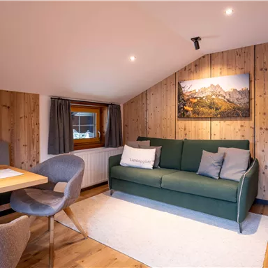Ein gemütlicher Wohnraum mit einem grünen Sofa und einem Essbereich. Die Wände sind mit Holz verkleidet und ein Bild einer Berglandschaft hängt an der Wand.