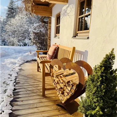 Ein gemütlicher Holzbank mit einem Herz-Motiv, das Holz für den Kamin lagert. Der Weg ist von frischem Schnee umgeben, und im Hintergrund sind schneebedeckte Bäume zu sehen.