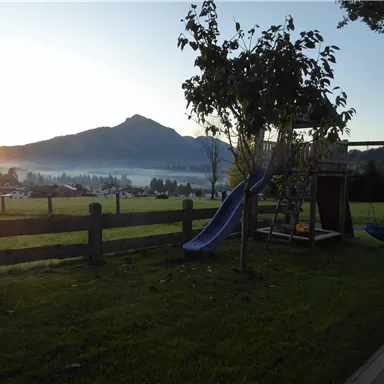 Ein schöner Garten mit einer Rutsche und einem Spielplatz. Im Hintergrund sind Berge und ein Sonnenaufgang sichtbar.