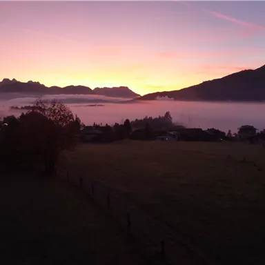 Eine malerische Landschaft bei Sonnenaufgang mit Bergen im Hintergrund. Der Himmel ist in sanften Farben gefärbt, während Nebel über das Tal zieht.