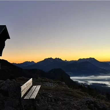 A bench is situated on a mountainside overlooking the mountains at sunrise. The sky is in warm colors, and fog drifts over the valley.