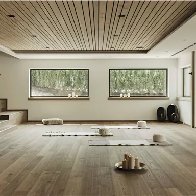 Ein ruhiger Raum mit Holzboden, großen Fenstern und einer minimalistischen Einrichtung. Auf dem Boden liegen Matten und Kissen für Entspannungsübungen.