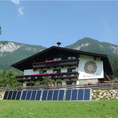A cozy, traditional farmhouse in the mountains. Solar panels are installed in the foreground to harness eco-friendly energy.