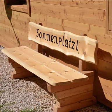 A wooden bench with the inscription "Sonnenplatzl". It stands in front of a wooden paneling and is surrounded by gravel.