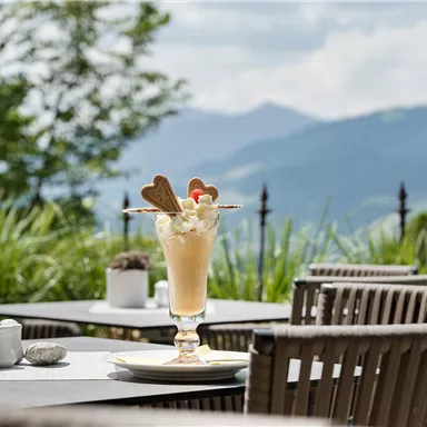 Ein köstliches Dessert in einem eleganten Glas steht auf einem Tisch im Freien. Im Hintergrund sind grüne Berge und ein blauer Himmel zu sehen.