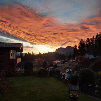 Ein wunderschöner Sonnenuntergang mit leuchtenden Farben über einer malerischen Landschaft. Im Vordergrund sieht man einen gepflegten Garten und Häuser im Hintergrund.