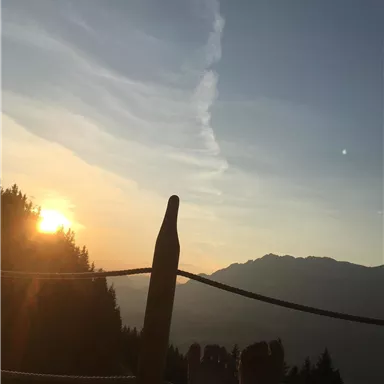 Ein Sonnenuntergang über den Bergen mit Blick auf einen klaren Himmel. Im Vordergrund sind Füße zu sehen, die entspannt auf einer Terrasse ruhen.