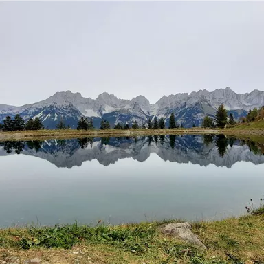 Ein ruhiger See spiegelt die umliegenden Berge wider. Die Landschaft ist von sanften grünen Wiesen umgeben.