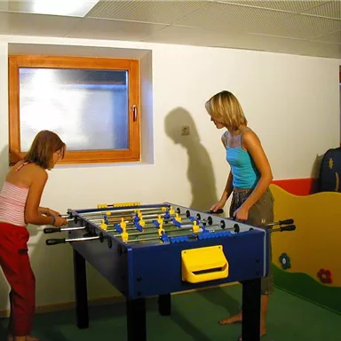 Two children are playing foosball in a bright room. Colorful furniture can be seen in the background.