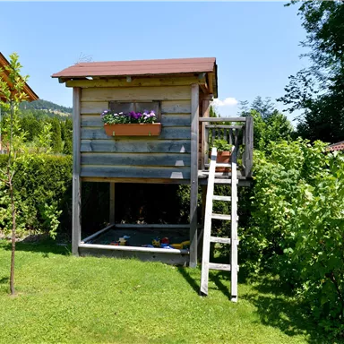 Ein hübsches Spielhaus aus Holz mit einer Treppe und einem Blumenkasten. Umgeben von einem grünen Garten und Bäumen.