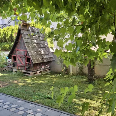 Ein kleines, traditionelles Gartenhaus mit einem schrägen Dach steht in einer grünen Wiese. Umgeben von Pflanzen und Bäumen bietet es eine gemütliche Atmosphäre.