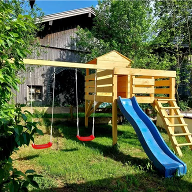 Ein Spielplatz mit einer Rutsche und zwei Schaukeln steht im grünen Garten. Im Hintergrund sieht man eine Holzstruktur und viel frisches Laub.