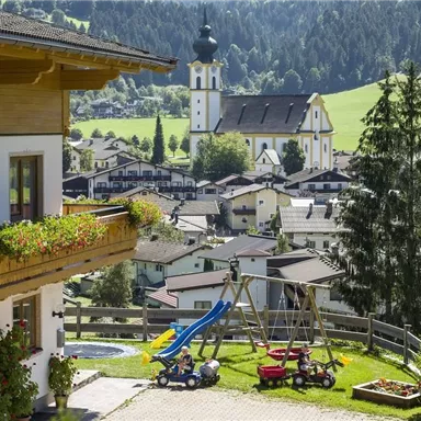 Eine malerische Landschaft mit einem traditionellen Gebäude und einer Kirche im Hintergrund. Im Vordergrund befinden sich ein Spielplatz und bunte Spielzeuge auf einer Wiese.