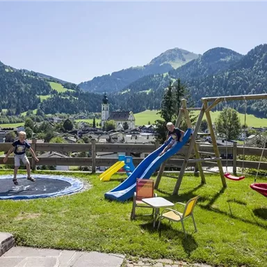 Eine schöne Landschaft mit Bergen im Hintergrund. Zwei Kinder spielen mit einem Trampolin und einer Rutsche im Garten.