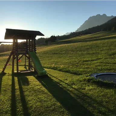 Ein Spielplatz mit einer Rutsche und Schaukeln steht auf einer grünen Wiese. Im Hintergrund sind Berge und ein klarer Himmel zu sehen.