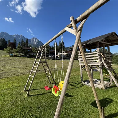 Ein Spielplatz mit einer Holzschaukel und einer Rutsche auf der Wiese. Im Hintergrund sind Berge und ein klarer Himmel zu sehen.